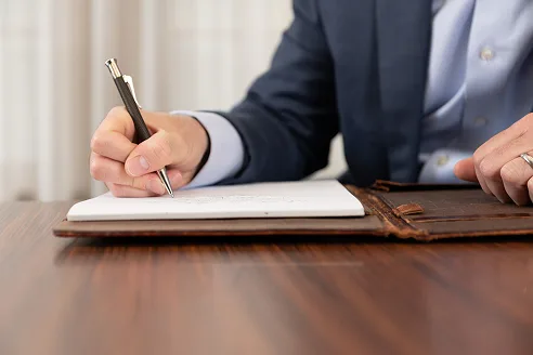 A man in a navy suit writing with a pen in a notebook inside a leather portfolio on a dark wood desk.